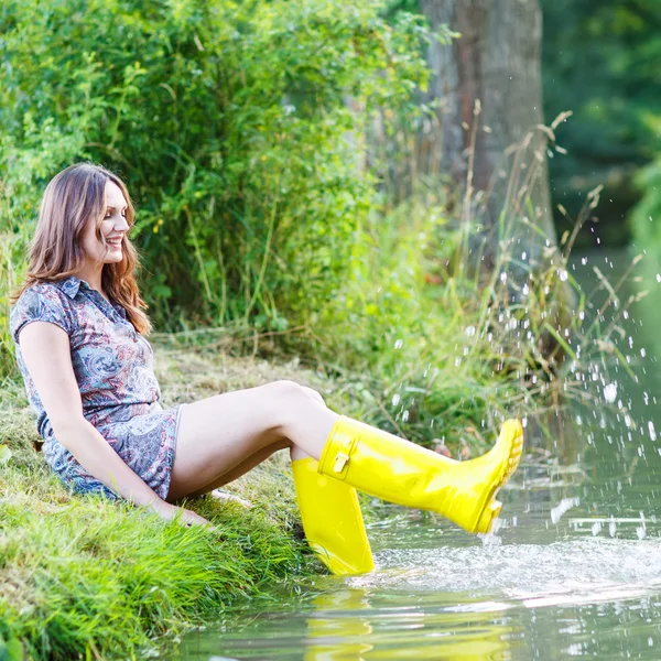 Jeune belle femme assise dans des bateaux en caoutchouc de pluie jaune par un r — Photo