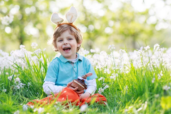 Милый счастливый маленький мальчик с пасхальными кроличьими ушами и поедающий шоколад. — стоковое фото