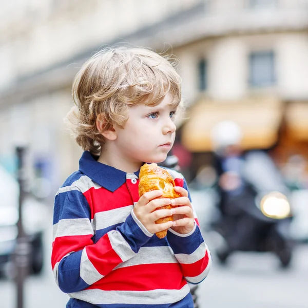 Pequena criança bonita em uma rua da cidade comendo croissant fresco — Fotografia de Stock