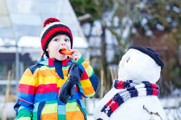 Lustige Junge in bunten Kleidern, die einen Schneemann, im Freien — Stockfoto