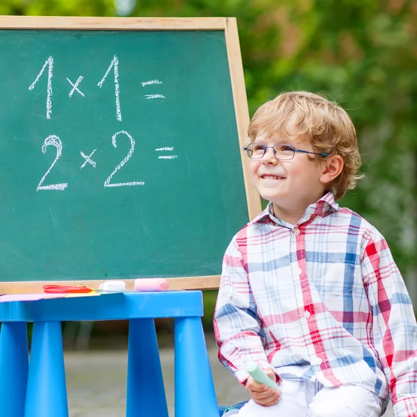 Симпатичный маленький мальчик в очках за школьной доской практикует матем — стоковое фото
