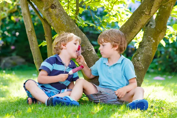 Two little sibling boys eating red ice cream in home's garden. — Stock Photo, Image