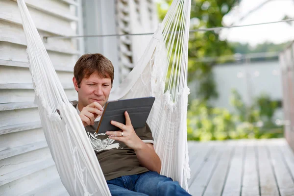 Hombre joven que relaja en la hamaca y usando la computadora de la tableta —  Fotos de Stock