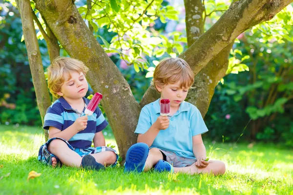 Deux petits frères et sœurs mangeant de la glace rouge dans le jardin de la maison . — Photo