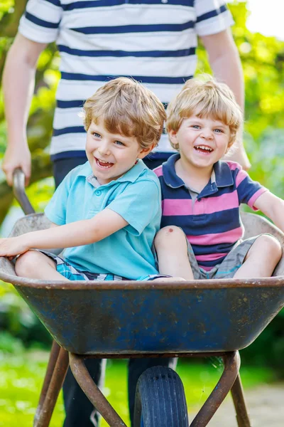 Two little boys having fun in wheelbarrow pushing by father — Stock Photo, Image