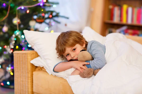 Cute little blond boy in his bed near Christmas tree with lights — Stock Photo, Image