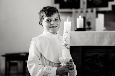 Küçük çocuk ilk kutsal komünyonunu alıyor. Vaftiz mumtutan mutlu çocuk. Katolik curch geleneği. Sunak yakınındaki bir kilisede beyaz geleneksel elbiseli çocuk