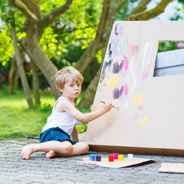 Kleiner Junge bemalt großes Papierhaus mit buntem Malkasten — Stockfoto