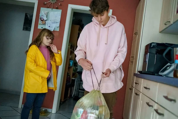 Genç çocuk kutuya plastik çöp getiriyor. Küçük kız kardeş kız kardeşine yardım ediyor. Çocuklar için ev yardımı