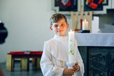Küçük çocuk ilk kutsal komünyonunu alıyor. Vaftiz mumtutan mutlu çocuk. Katolik curch geleneği. Sunak yakınındaki bir kilisede beyaz geleneksel elbiseli çocuk