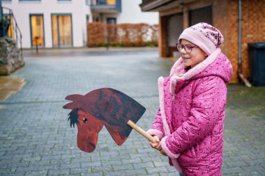 Pembe ceketli gülümseyen küçük bir kız, sakin bir yerleşim yerinde el yapımı ahşap bir atla dışarıda oynar. Çocukluk anıları, gerçek hayat sahneleri.