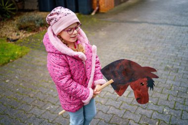 Pembe ceketli gülümseyen küçük bir kız, sakin bir yerleşim yerinde el yapımı ahşap bir atla dışarıda oynar. Çocukluk anıları, gerçek hayat sahneleri.
