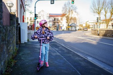 Pembe kask takan küçük liseli kız, sakin bir kaldırım boyunca scooter sürüyor. Gerçek hayatta günlük hareketlilik, bağımsızlık ve güvenliği gösteriyor..