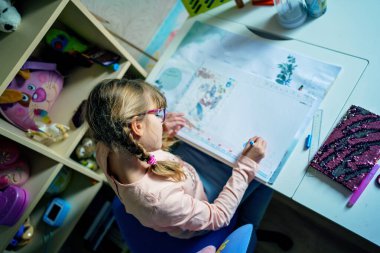 Gözlüklü genç bir kız evdeki bir masada oturuyor, rahat bir çocuk odasında çiziyor ve öğreniyor, doğal ışık, gerçek günlük aile hayatı ve eğitim sahnesi.