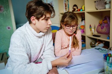 Büyük kardeş ergen oğlan, çocuk odasında bir masada ödevi olan genç bir kıza yardım ediyor, evde sessiz bir öğrenme anı, gerçek aile hayatı, doğal ışık, günlük eğitim sahnesi..