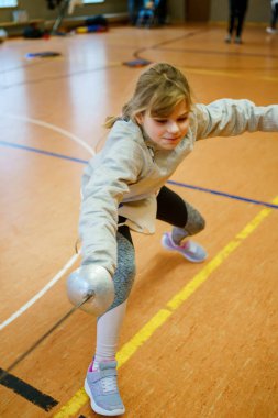 Genç kız kapalı alanda eskrim antrenmanı yapıyor. Spor salonundaki antrenman sırasında odaklanmış ve kendinden emin. Koruyucu ekipmanlarla gerçek bir çocukluk aktivitesi.