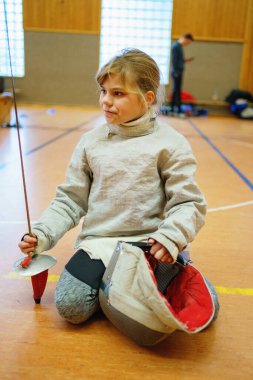 Genç kız kapalı alanda eskrim antrenmanı yapıyor. Spor salonundaki antrenman sırasında odaklanmış ve kendinden emin. Koruyucu ekipmanlarla gerçek bir çocukluk aktivitesi.