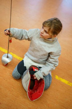 Genç kız kapalı alanda eskrim antrenmanı yapıyor. Spor salonundaki antrenman sırasında odaklanmış ve kendinden emin. Koruyucu ekipmanlarla gerçek bir çocukluk aktivitesi.