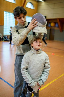 Genç çocuk, genç kızın spor salonunda eskrim maskesi takmasına, antrenman sırasında destek olmasına, gerçek insanlarla gerçek hayatta spor yapmasına yardım ediyor.