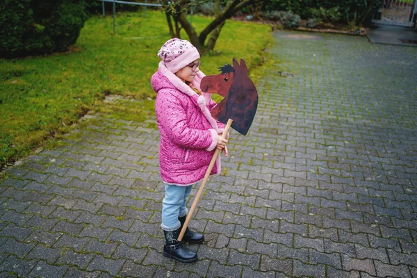 Pembe ceketli gülümseyen küçük bir kız, sakin bir yerleşim yerinde el yapımı ahşap bir atla dışarıda oynar. Çocukluk anıları, gerçek hayat sahneleri.