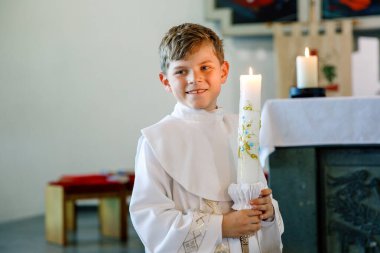 Küçük çocuk ilk kutsal komünyonunu alıyor. Vaftiz mumtutan mutlu çocuk. Katolik curch geleneği. Sunak yakınındaki bir kilisede beyaz geleneksel elbiseli çocuk