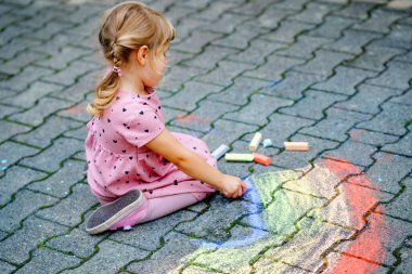 Küçük anaokulu kızı arka bahçede renkli tebeşirlerle gökkuşağını boyuyor. Çocuk resim çizerken ve resim çizerken çok mutlu. Yazın yaratıcı açık hava aktivitesi