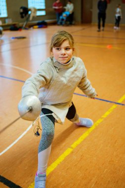 Genç kız kapalı alanda eskrim antrenmanı yapıyor. Spor salonundaki antrenman sırasında odaklanmış ve kendinden emin. Koruyucu ekipmanlarla gerçek bir çocukluk aktivitesi.