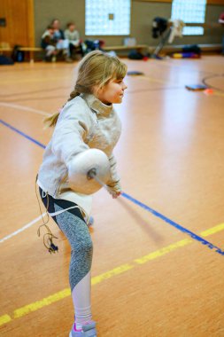 Genç kız kapalı alanda eskrim antrenmanı yapıyor. Spor salonundaki antrenman sırasında odaklanmış ve kendinden emin. Koruyucu ekipmanlarla gerçek bir çocukluk aktivitesi.