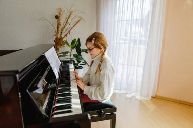 Gözlüklü gülümseyen kız evde piyano çalıyor, rahat bir kazak giyiyor, açık havada müzik çalışmasının tadını çıkarıyor..