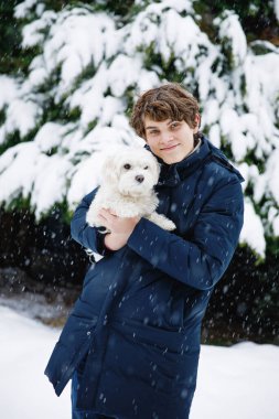 Genç bir çocuk, kar yağarken dışarıda küçük beyaz bir köpek tutuyor sıcak bir kışlık ceket giyiyor ve karlı bir bahçede hafifçe gülümsüyor..