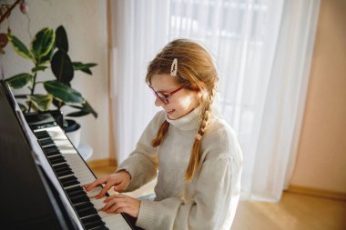 Gözlüklü gülümseyen kız evde piyano çalıyor, rahat bir kazak giyiyor, açık havada müzik çalışmasının tadını çıkarıyor..