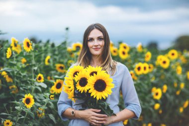 Ayçiçeği tarlasında elinde bir buket parlak sarı ayçiçeği tutan gülümseyen kadın, sakin bir yaz portresi, doğal gerçek hayat anı.