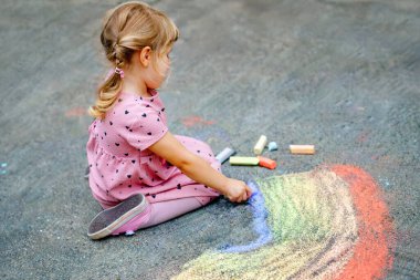 Küçük anaokulu kızı arka bahçede renkli tebeşirlerle gökkuşağını boyuyor. Çocuk resim çizerken ve resim çizerken çok mutlu. Yazın yaratıcı açık hava aktivitesi