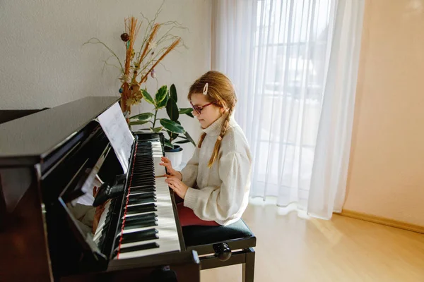 Gözlüklü gülümseyen kız evde piyano çalıyor, rahat bir kazak giyiyor, açık havada müzik çalışmasının tadını çıkarıyor..
