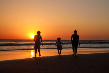 Gün batımında plajda koşup zıplayan üç çocuk silueti. Mutlu bir aile, iki okul çocuğu ve küçük bir anaokulu kızı. Kardeşler birlikte eğleniyor. Bağ kurmak ve aile tatili