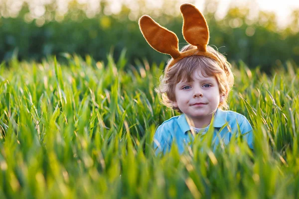 Cute blond kid boy with Easter bunny ears playing in green fieldon sunny spring day, celebrating Easter holiday — 图库照片