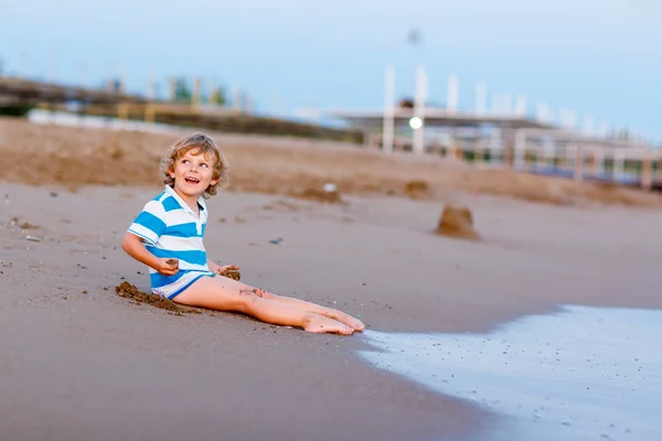 Menino pequeno feliz se divertindo com castelo de areia pelo oceano — Fotografia de Stock