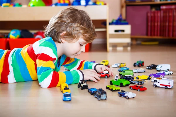 Маленький блондинка играет с большим количеством игрушечных автомобилей в помещении — стоковое фото