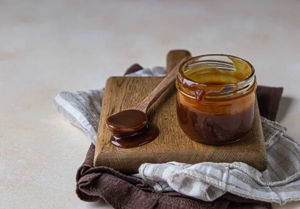 Salsa al caramello fatta in casa in un barattolo di vetro e cucchiaio su tavola di legno. Deliziosa salsa con sale marino per gelato e dessert. — Foto Stock