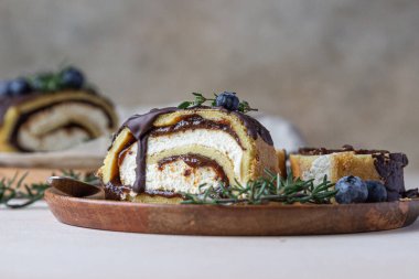 Çikolata kaplı krem peynirli sünger kek rulosu, parşömen üzerinde yaban mersini ve biberiye, açık beton arka plan. Bisküvi İsviçre böreği..