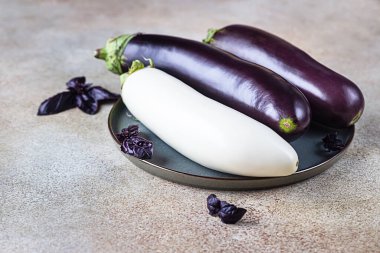 Taze çiğ, mor ve beyaz patlıcan tabakta ve arka planda mor fesleğen var..