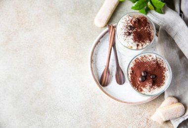 Geleneksel İtalyan tatlı tiramisusu, cam kaplı, hafif taşlı arka plan. Kadın parmaklarından yapılmış kahve aromalı porsiyon tatlı ve kakao tozuyla süslenmiş mascarpone. Üst görünüm.
