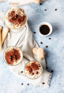Geleneksel İtalyan tatlı tiramisusu, cam kaplı, hafif taşlı arka plan. Kadın parmaklarından yapılmış kahve aromalı porsiyon tatlı ve kakao tozuyla süslenmiş mascarpone. Üst görünüm.