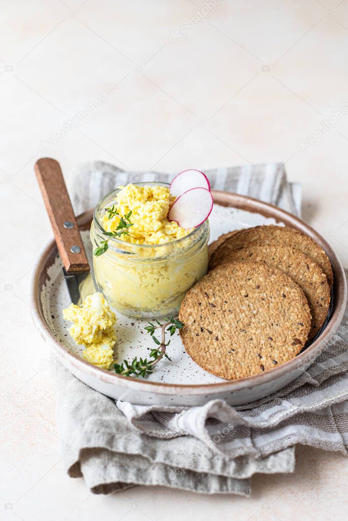 Egg pate, salad or dip in glass jar served with radish, thyme and multigrain crackers, light concrete background.