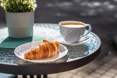 Kahvaltıda kruvasan ve bir fincan cappuccino, sabahın erken saatlerinde bir sokak kafesinde masanın üstünde. Seçici odak.