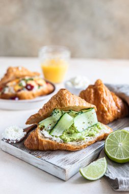 Ricotta ya da krem peynirli kruvasan sandviçi, ahşap kesim tahtasının üzerinde avokado püresi ve salatalık. Lezzetli bir kahvaltı ya da kahvaltı. Seçici odak.