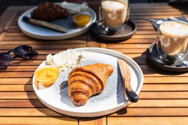 Açık kafedeki ahşap masada taze kruvasan, portakal reçeli ve tereyağlı kapuçino ile kahvaltı. Güneşli sabah.
