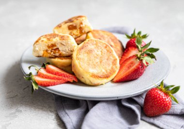 Süzme peynirli krep ya da çilekli börek ve açık gri taşlı arka planda doğal yoğurt. Sağlıklı kahvaltı ya da öğle yemeği. Syrniki..