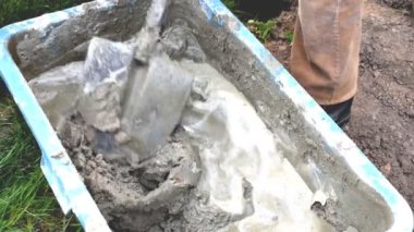 Eldivenleri karıştıran, çimento çamuru karıştıran, beton harcı büyük kasede kürekle karıştıran işçi elleri, su deposu, yalak, kova. İnşaat süreci, yenileme, temel atma hazırlıkları.