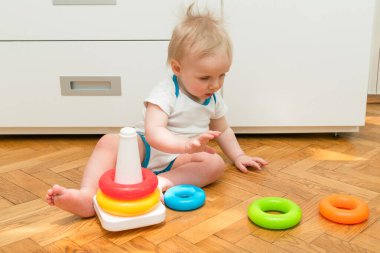 Evde renkli piramit oyuncağı oynayan sevimli, beyaz, bebek bir çocuk. Mutlu çocuk plastik halkalardan kule inşa ederken eğleniyor. Çocukların mantık gelişimi ve mutlu çocukluk kavramı.